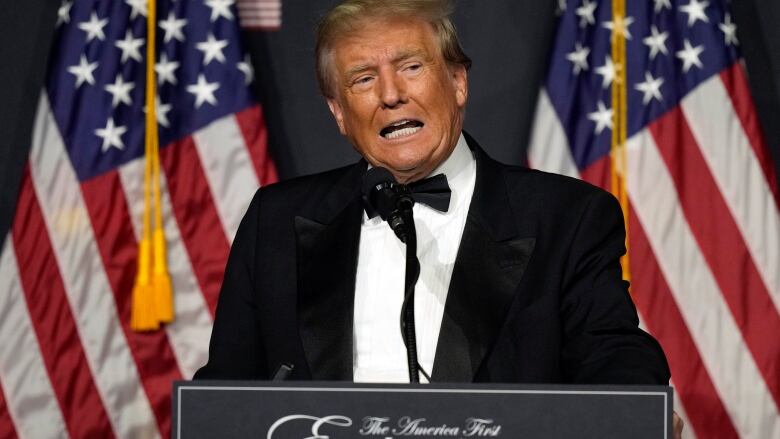 Man in a tuxedo standing at a podium in front of American flags.