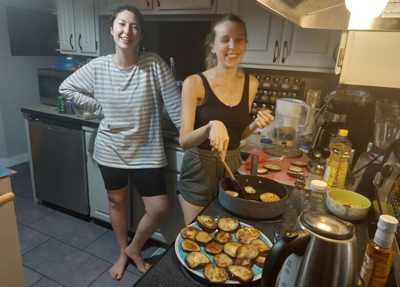 Two young women stand in a kitchen. One is frying something that looks like zucchini slices on the stove. 