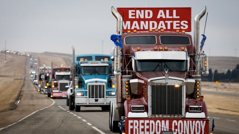 A line of semis can be seen on the highway, driving away from the protest. On top of the first truck sits a red and white sign reading 