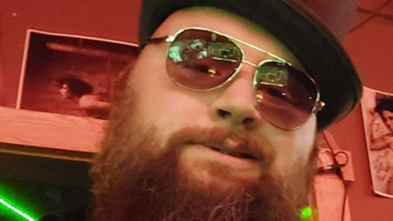 A man with a bushy beard wearing sunglasses and a hat with several posters on a wall in the background. 