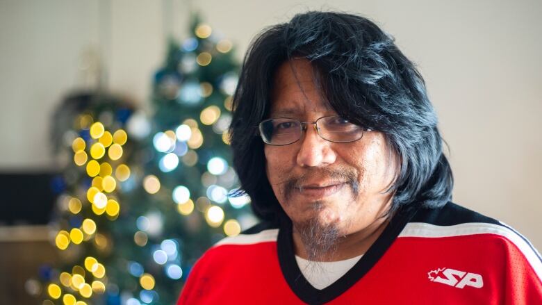 A man with thick black hair, glasses and a goatee stands in front of Christmas trees.