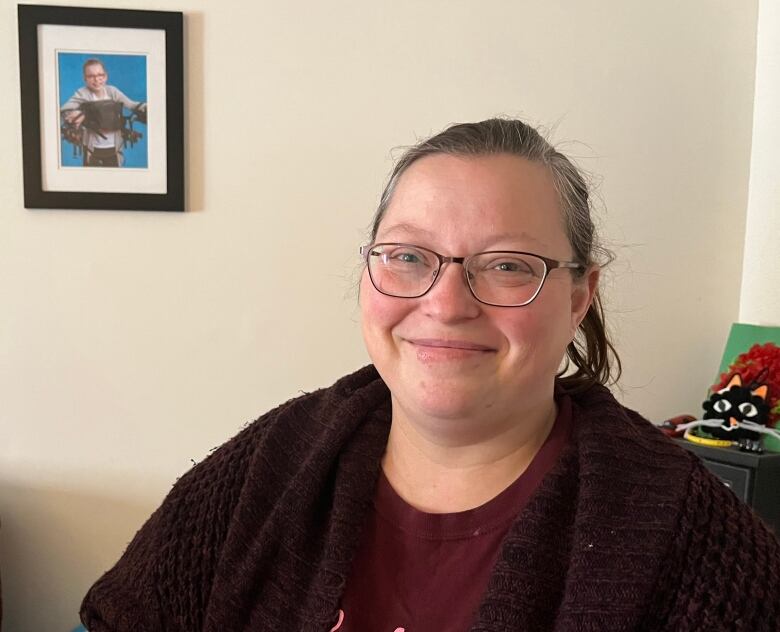 A middle-aged woman smiles into the camera. Behind her, on the left, a framed photo on the wall shows her daughter Claire.