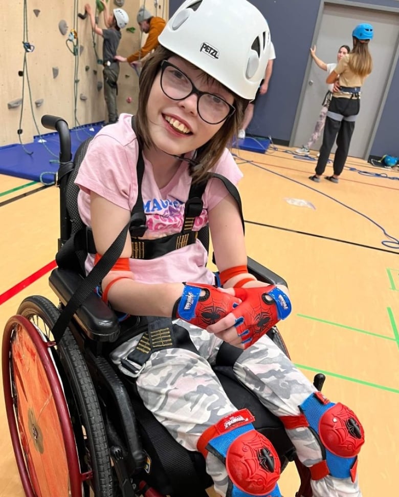 A girl sits in a wheelchair and smiles into the camera. She wears a helmet as well as protective gear on her hands and knees. There's an artificial rock wall behind her, with two people standing in front of it.