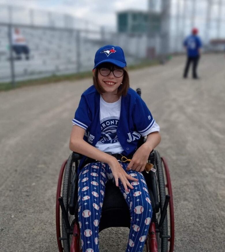 A young girl smiles into the camera. She sits in a wheelchair and wears baseball fan attire from head to toe. In the background, there are baseball spectator ranks.