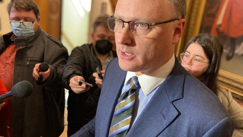 Bald man with glasses stands in lobby of legislature with reporters around him.