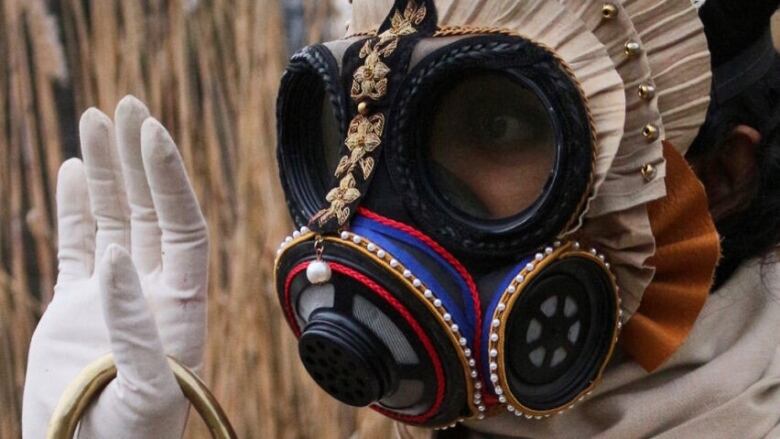 Crop of a photograph by artist Rajni Perera. Depicts a close-up of a brown woman wearing a decorated gas mask, photographed outdoors with tall grass in the background.