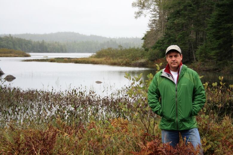 A man stands in the foreground with water and forest behind him.