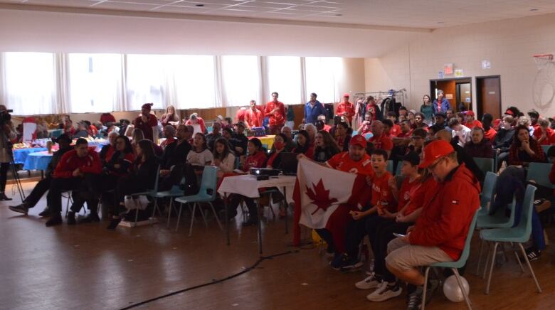 Fans sit in chairs in a church hall, most wearing red Canadian shirts and jerseys, including a father and son with a flag in the front row