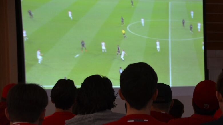 A row of heads sit in the dark in front of a large screen showing a soccer game 