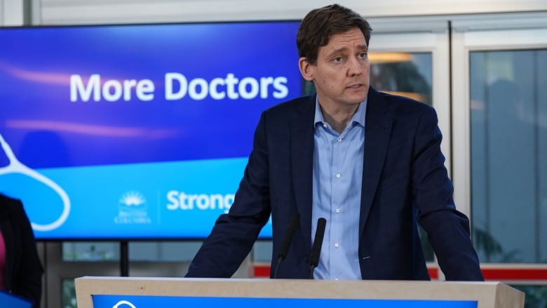 David Eby speaks at a podium that reads 'More Doctors'.