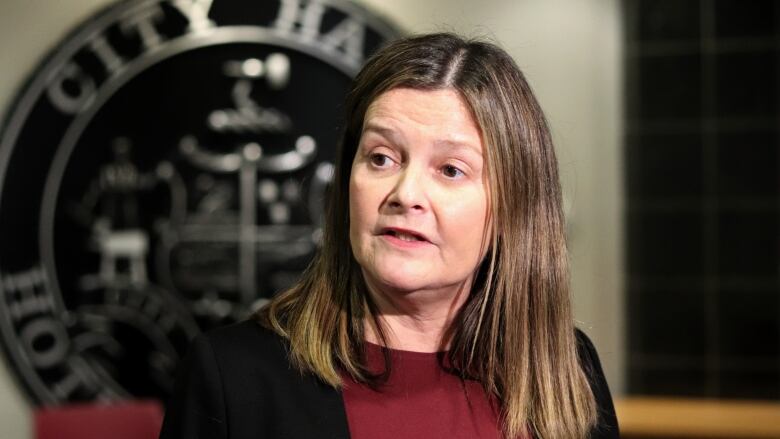 A woman with shoulder-length brown hair wearing a red shirt and black jacket in front of a large Moncton logo that says 