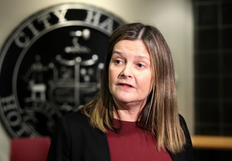 A woman with shoulder-length brown hair wearing a red shirt and black jacket in front of a large Moncton logo that says 