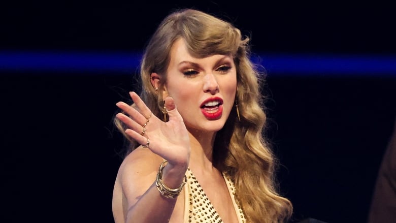 Taylor Swift, a blonde woman wearing a gold dress, waves at an awards ceremony.