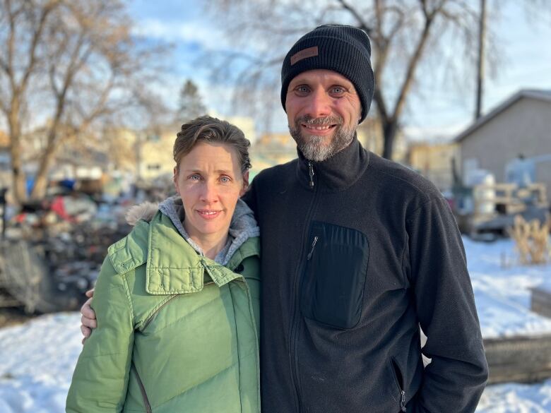 A man and a woman are standing next to each other. Behind them, debris can be seen strewn across a property with no house on it.