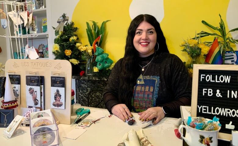 A woman wearing a black shirt sits at a desk in front of a yellow wall.