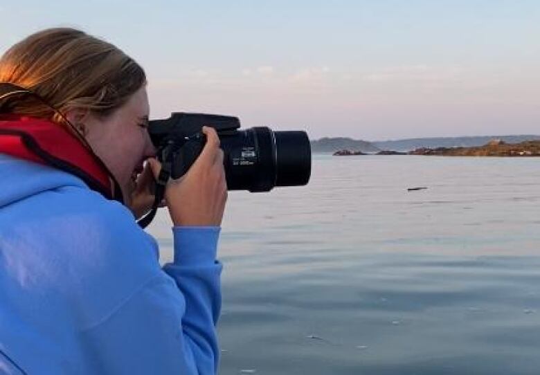 A woman in a bright blue jacket leans over the edge of a boat and peers through a camera with a long lens. 