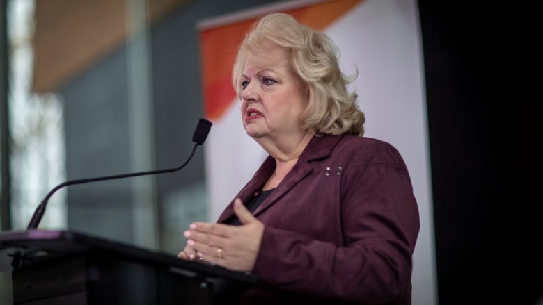 A woman stands at a podium gesturing in mid-speech.