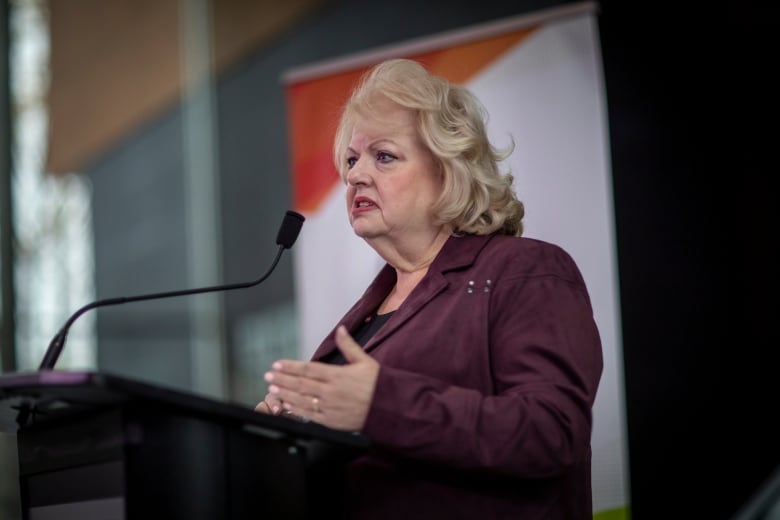 A woman stands at a podium gesturing in mid-speech.