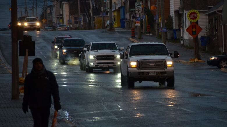 Cars drive through the dark along a city street lined with telephone polls, with a man walking on the sidewalk in the foreground. 