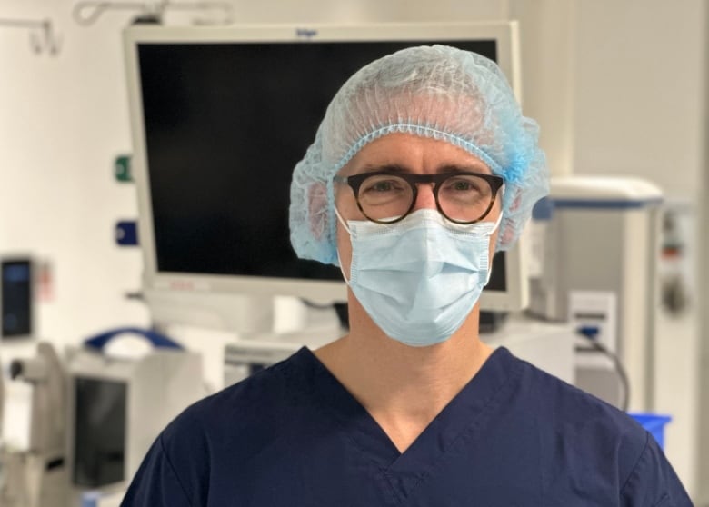 A doctor in a mask, glasses and scrubs stands in an operating room.