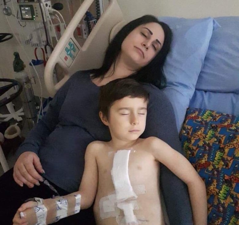 A child lies sleeping in a hospital bed, with an oxygen tube attached to his nose. His mother lies beside him, looking at the camera.