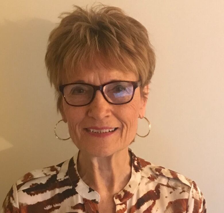 A woman with short hair, glasses, hoop earrings and wearing a patterned blouse. We see her from the shoulders up. 