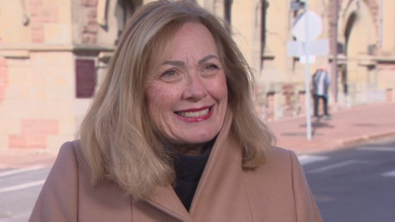 A white woman with a blonde bob, red lipstick and a beige coat stands outside on a street