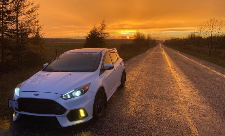 A white car sits on a two-lane road running through fields as the sun rises.