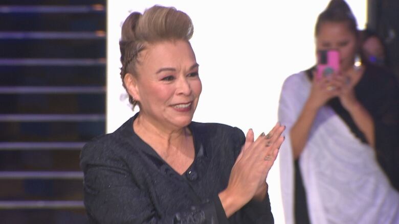 Pam Baker at the end of her ready-to-wear collection showing, wears an elegant black suit and is clapping along with the audience as she walks down the runway, smiling.