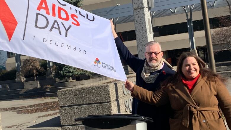 A man and a woman in winter jackets pose with a World Aids Day flag. 