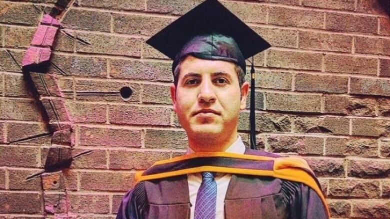 Ayub Khezrnejad stands in front of a grey brick wall wearing a graduation gown, holding his Master's of Engineering diploma from Memorial University.