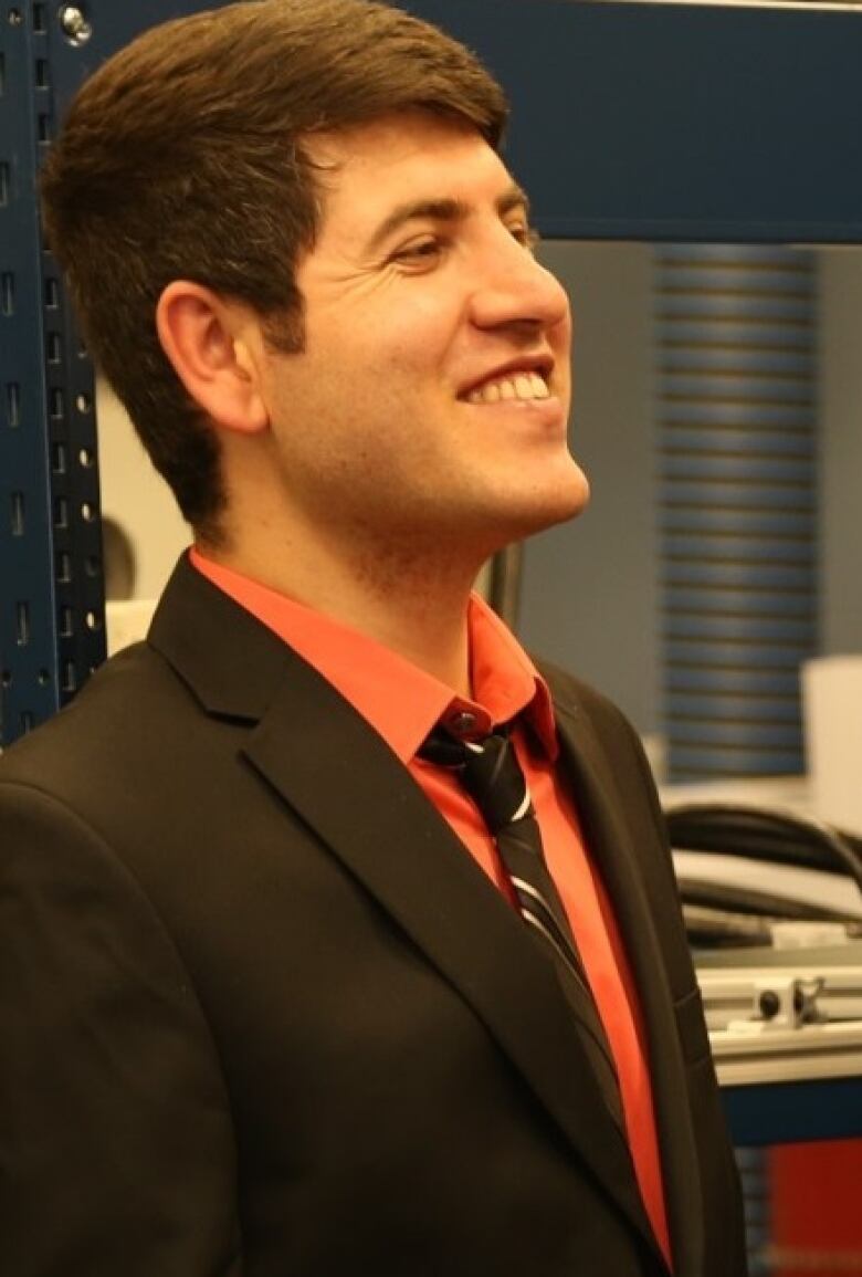 A man stands side on looking to the right. He is wearing a black suit jacket, a burnt orange button-up shirt, and a black and white striped tie. He is smiling.