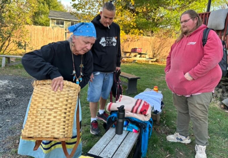 Elder Imelda Perley stands outside next to a bench, holding her basket. Two men, one in jean shorts and a black hoodie and one in green pants and a red sweater, stand with her.