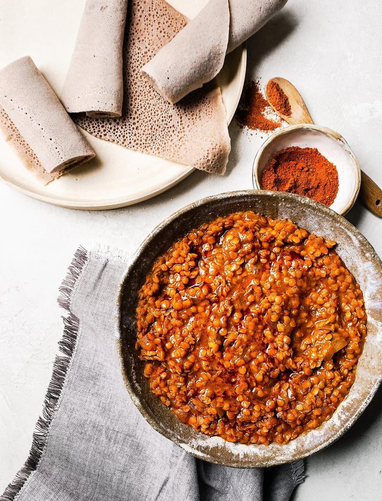 overhead shot of a bowl of lentil stew with a small bowl of spices sitting above it. a plate with flatbread (injera) sits next to it. 