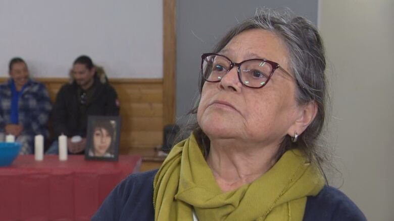 A woman with grey hair, wearing glasses and a green scarf, looks at someone off-camera.