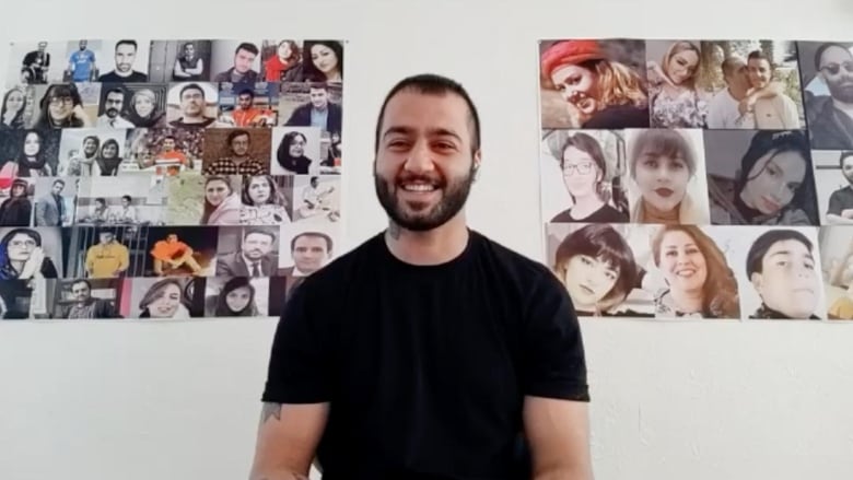 A short-haired and bearded man in a black shirt smiles in front of photo collages of people's faces.