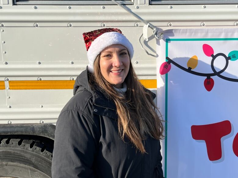 A woman with long brown hair wearing a black jacket and a sparkly red Santa hat stands in front of a white school bus.