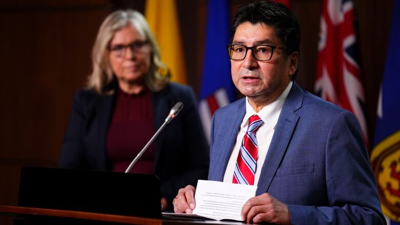 A man wearing a blue suit speaks at a podium with a woman standing behind.