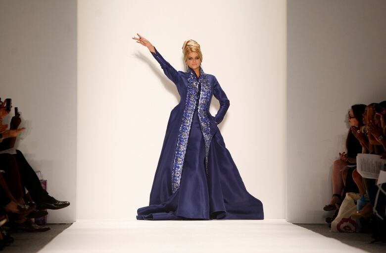 Individual in a long purple gown on a runway, arm raised in the air.