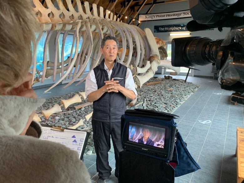 A man clasps his hands while standing in front of a humpback whale skeleton. In the foreground is a video camera recording an interview. 