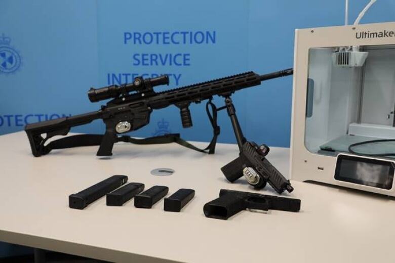 A black rifle sits on a table, along with ammunition clips, a pistol and a grey square-shaped printer.