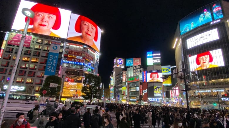 Crowds fill a public square, with bright billboards overhead.