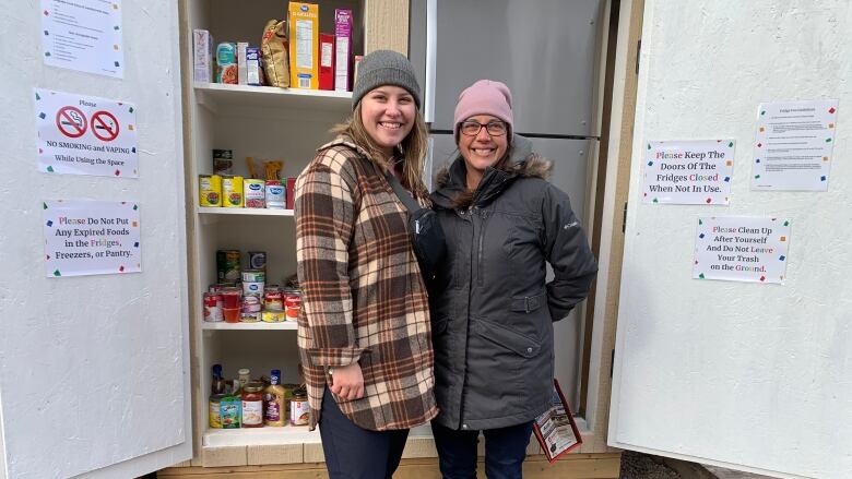 Two women stand in front of a fridge full of food 