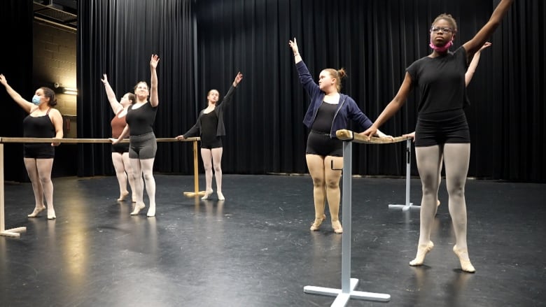 A group of dancers lean on a ballet bar and reach up to the sky. 