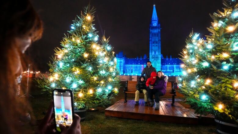 People pose for a smartphone photo between two Christmas trees with the Peace Tower on Parliament Hill in the back, lit in blue.