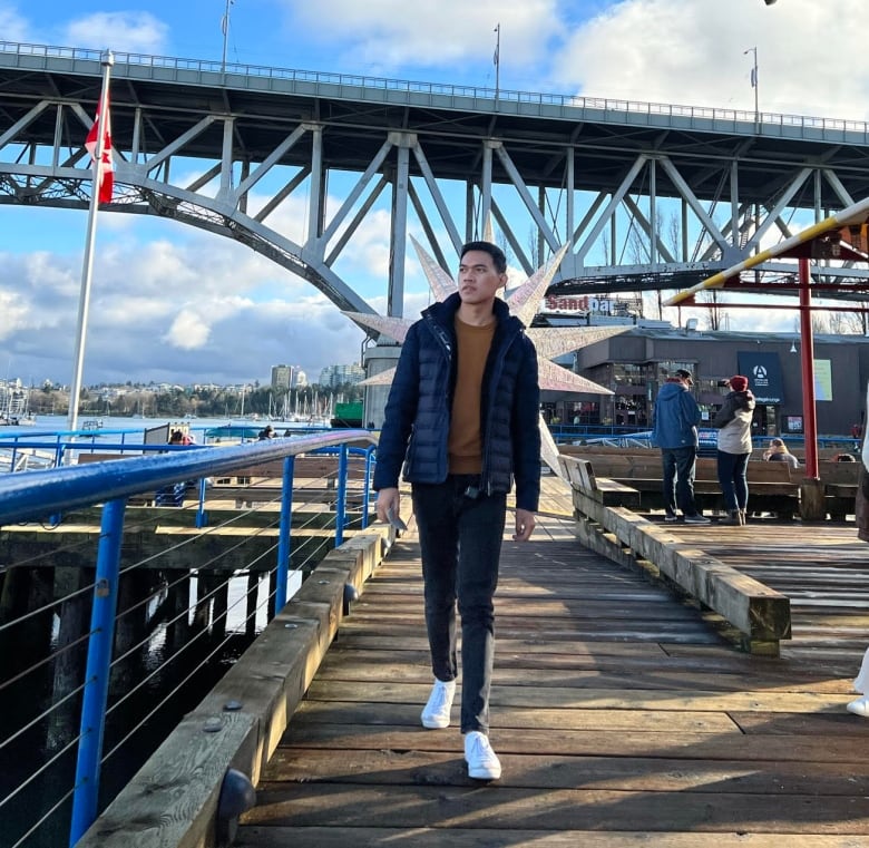 A man stands on a wooden bridge deck with a bridge in the background.