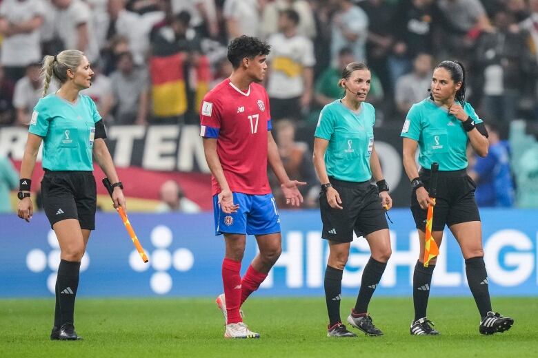 Three women in officals' uniforms walk across a soccer field, on either side of a man in a soccer player's uniform.