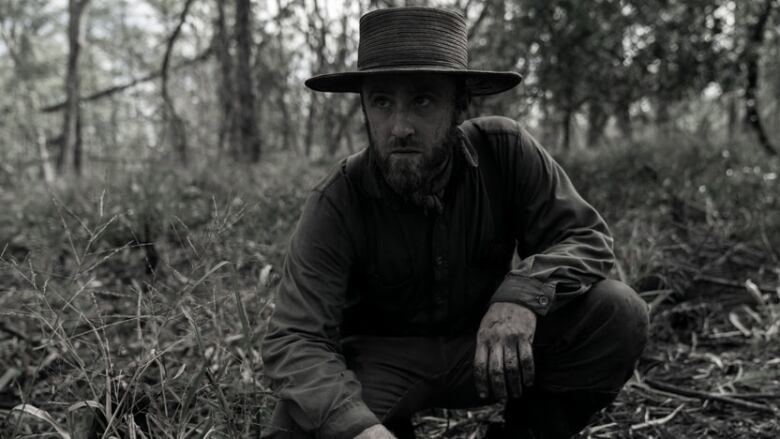 A man with a straw hat and plain-looking clothes kneels down. He has a rough looking beard and somewhat dirty hands. He is in a forest. 