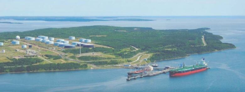 An aerial shot shows an oil and gas storage facility in the distance, with ships on the water in the foreground.