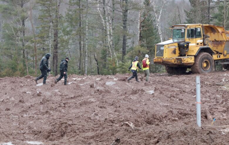 Four people are seen walking through a muddy field at a construction site. A large loader vehicle is seen in the background.